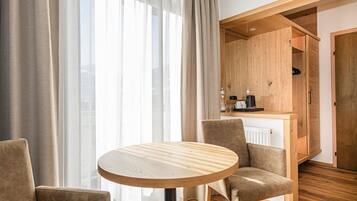 A modern room with a round wooden table, two chairs, a kitchen area with cabinets, and a large window with curtains.