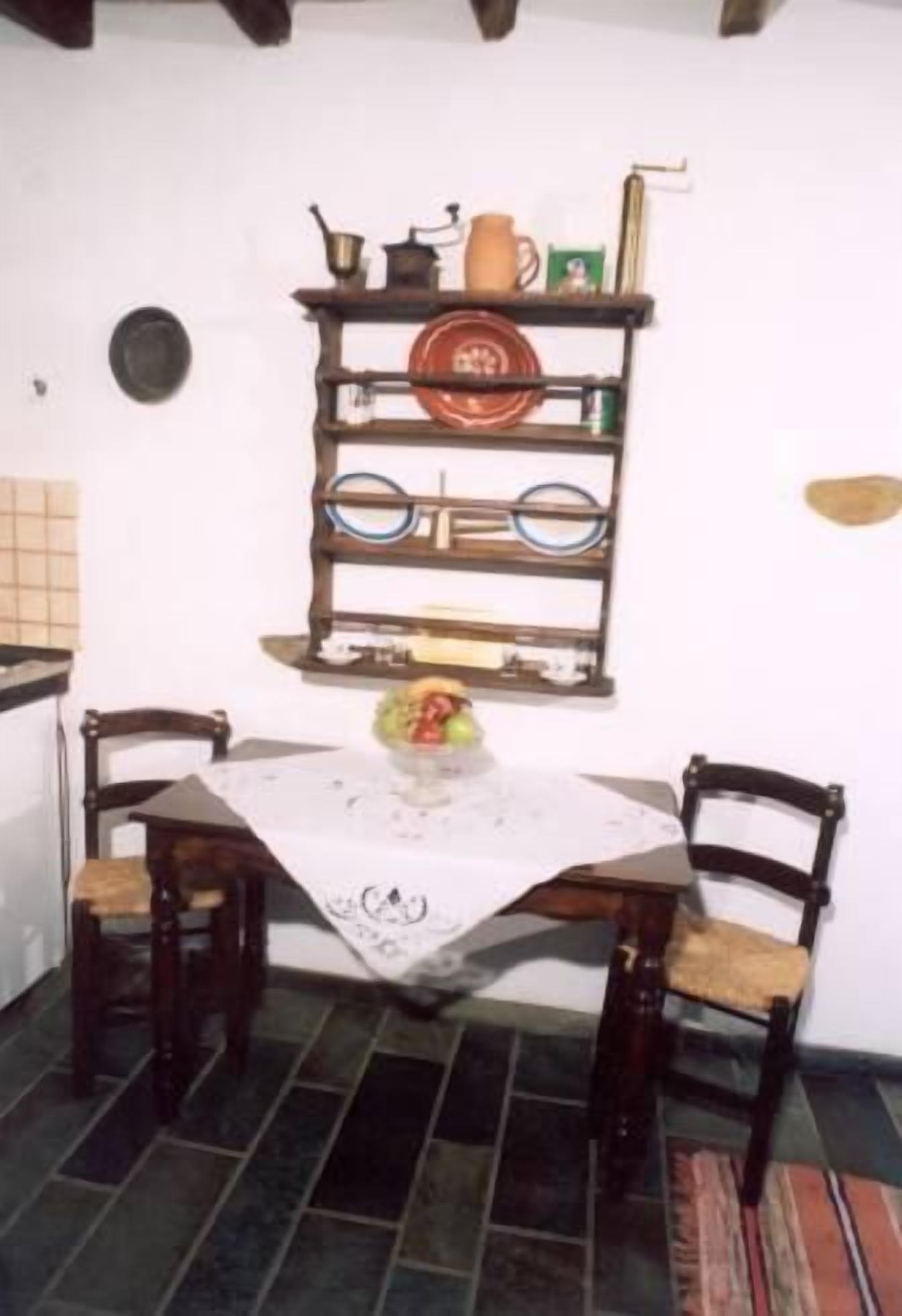 Un comedor con una mesa lista para dos personas, rodeada de sillas. Un estante sobre la mesa contiene distintos utensilios de cocina.