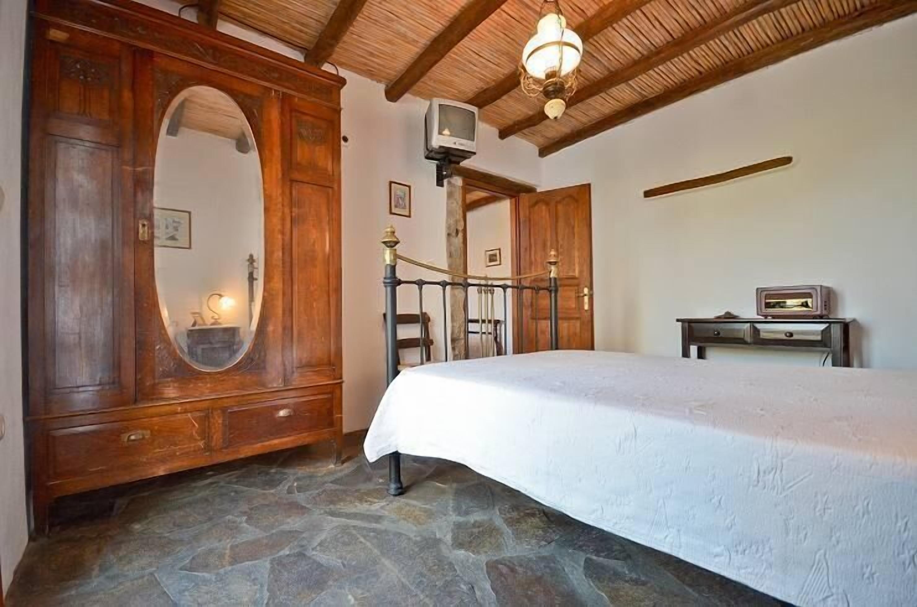 Une chambre avec un sol en pierre, des meubles en bois, un lit recouvert d’une courtepointe blanche, une armoire en bois, un miroir, une télévision et une petite table.