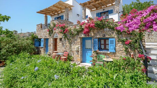 Una casa de piedra tradicional con contraventanas azules y pérgolas de madera, rodeada de abundante vegetación y flores coloridas.