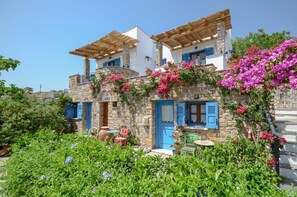 Ein traditionelles Steinhaus mit blauen Fensterläden und hölzernen Pergolen, umgeben von üppigem Grün und leuchtenden Blumen.