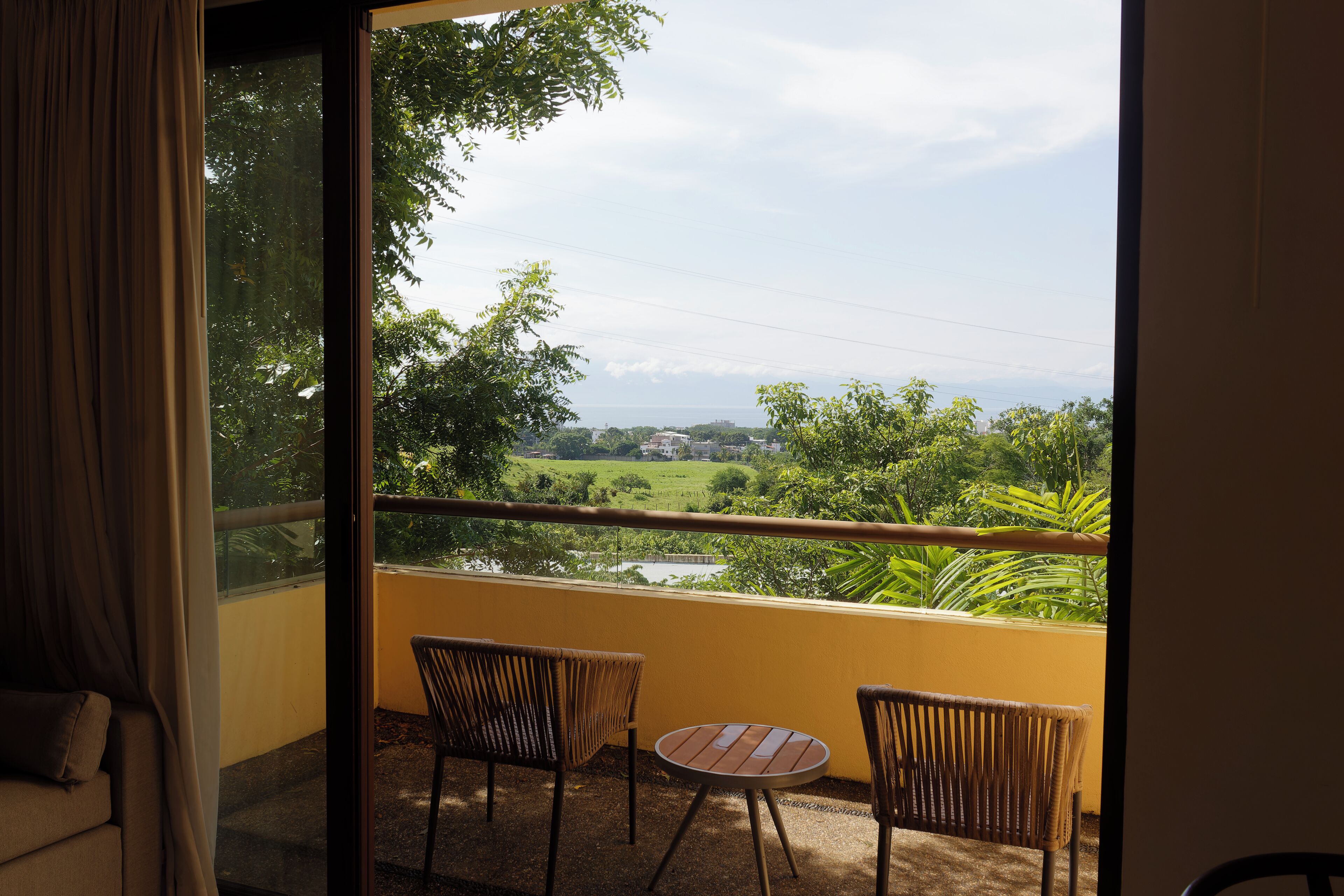 Un balcone con due sedie in vimini e un tavolino, affacciato su un paesaggio verde e un corpo d'acqua.