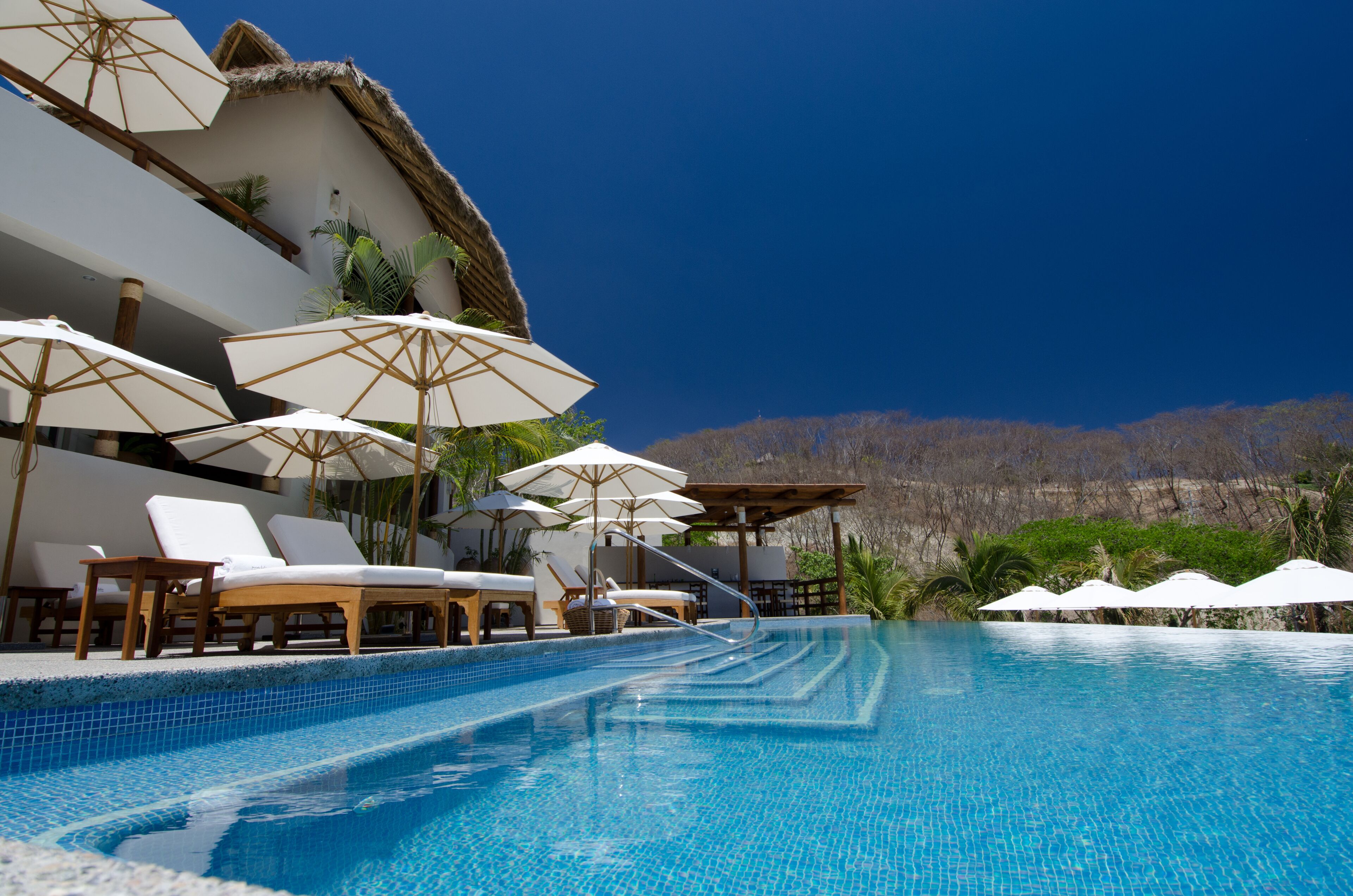 Zona junto a la piscina con sillones de descanso y sombrillas, piscina azul cristalina y montañas al fondo.