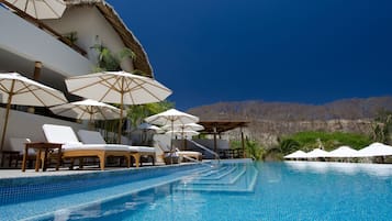 Zona junto a la piscina con sillones de descanso y sombrillas, piscina azul cristalina y montañas al fondo.