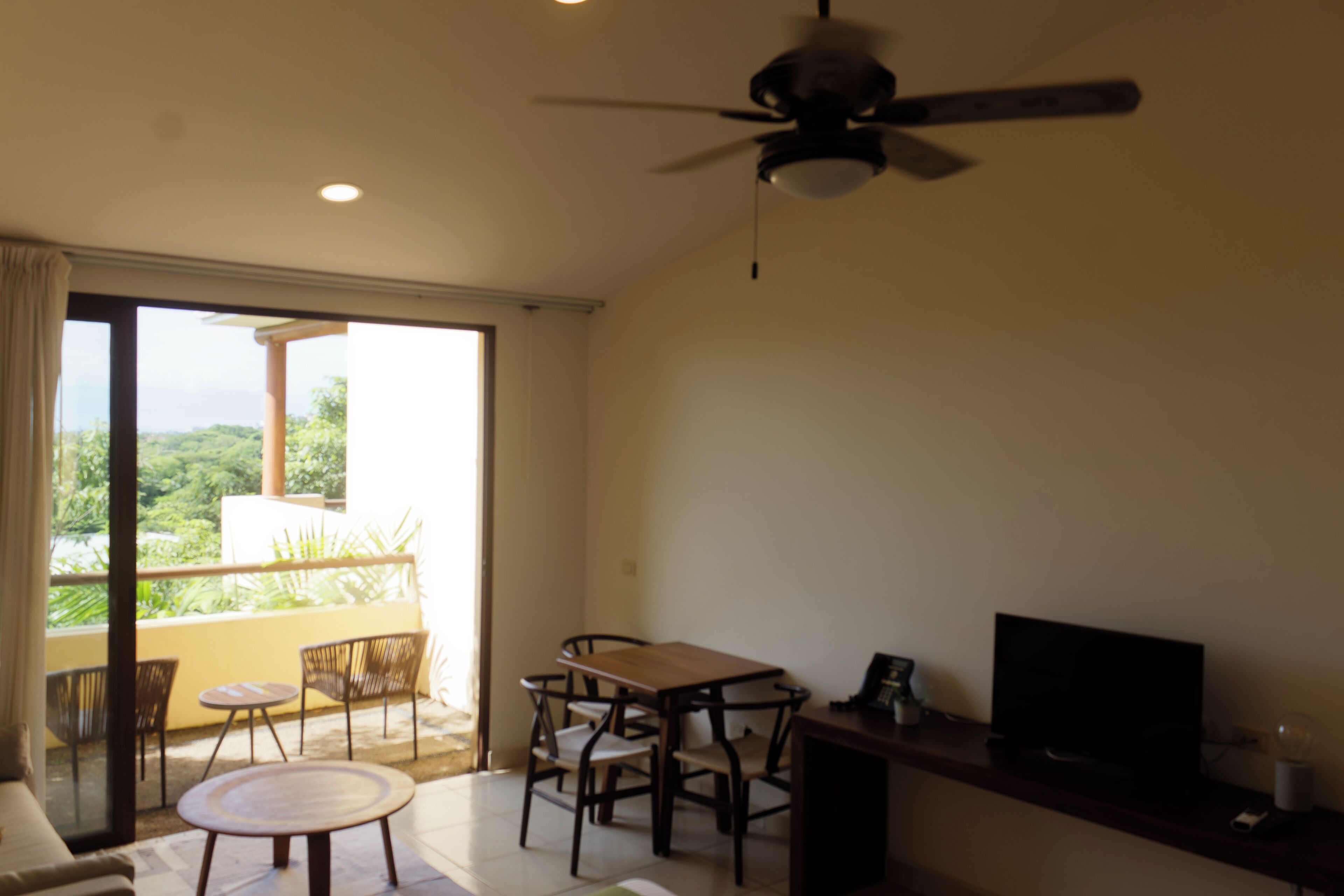 Una stanza con ventilatore a soffitto, televisore su supporto in legno, tavolo da pranzo con sedie e divano.
