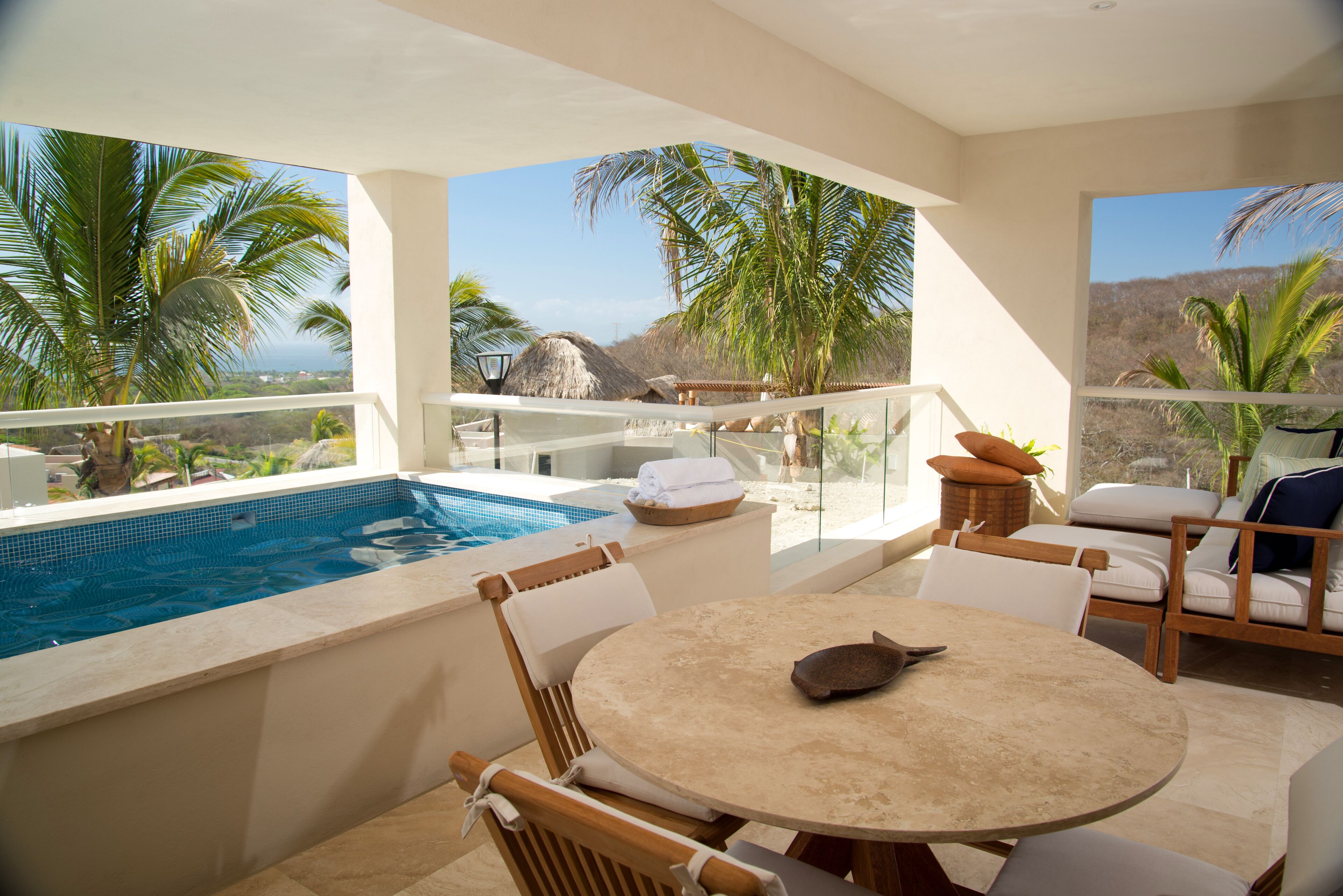 A poolside area with a table and chairs, a thatched-roof structure, and a view of palm trees and mountains.