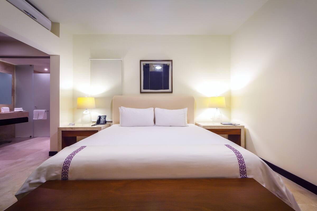 Habitación de hotel con una cama grande, dos mesitas de noche con lámparas, un espejo en la pared y suelo de madera.