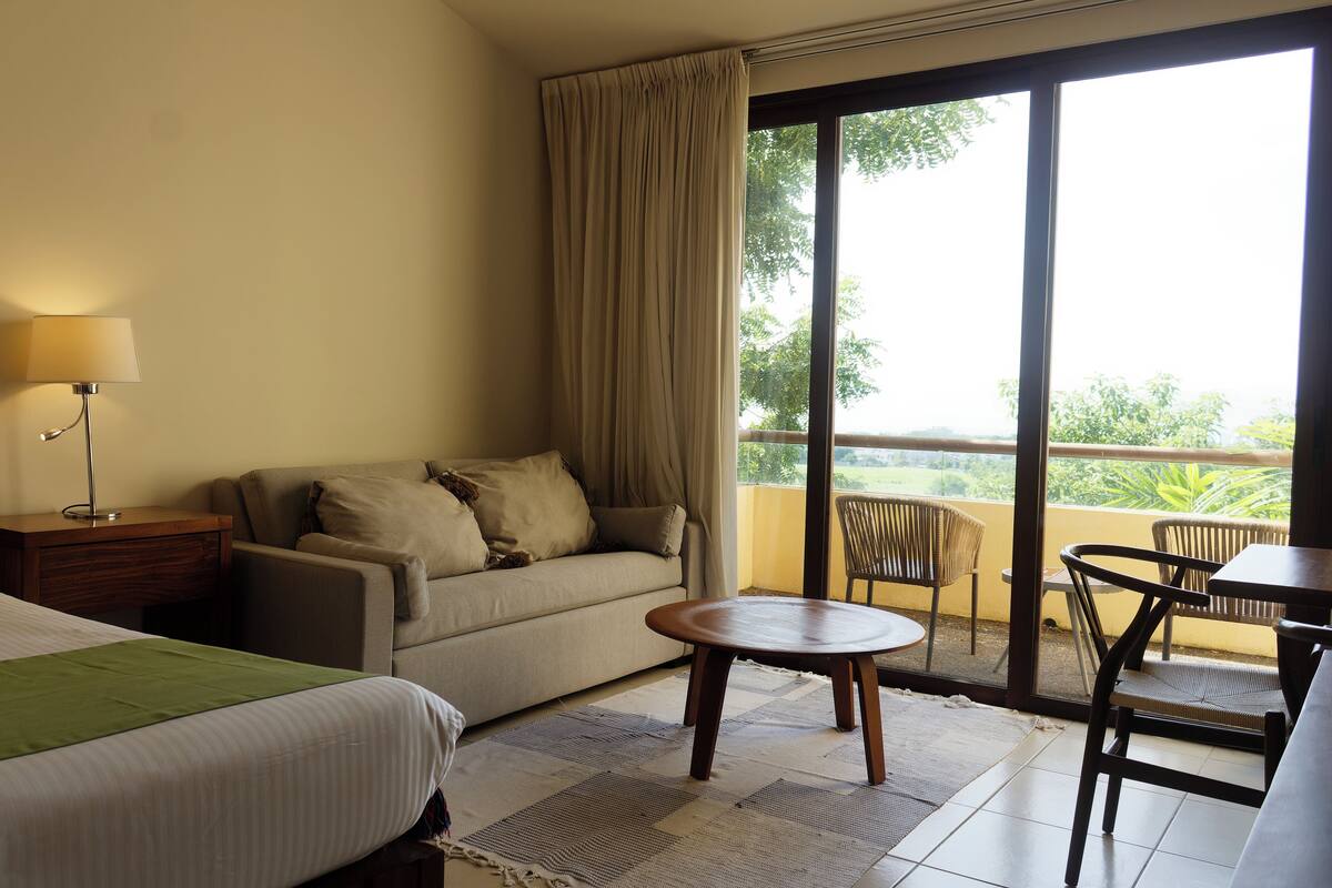 Habitación de hotel con cama, sofá, una mesita y balcón con sillas.