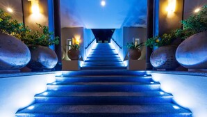 Eine Treppe mit blauer Beleuchtung, die zu einer höheren Etage führt, gesäumt von großen Kübelpflanzen und Wandleuchten.