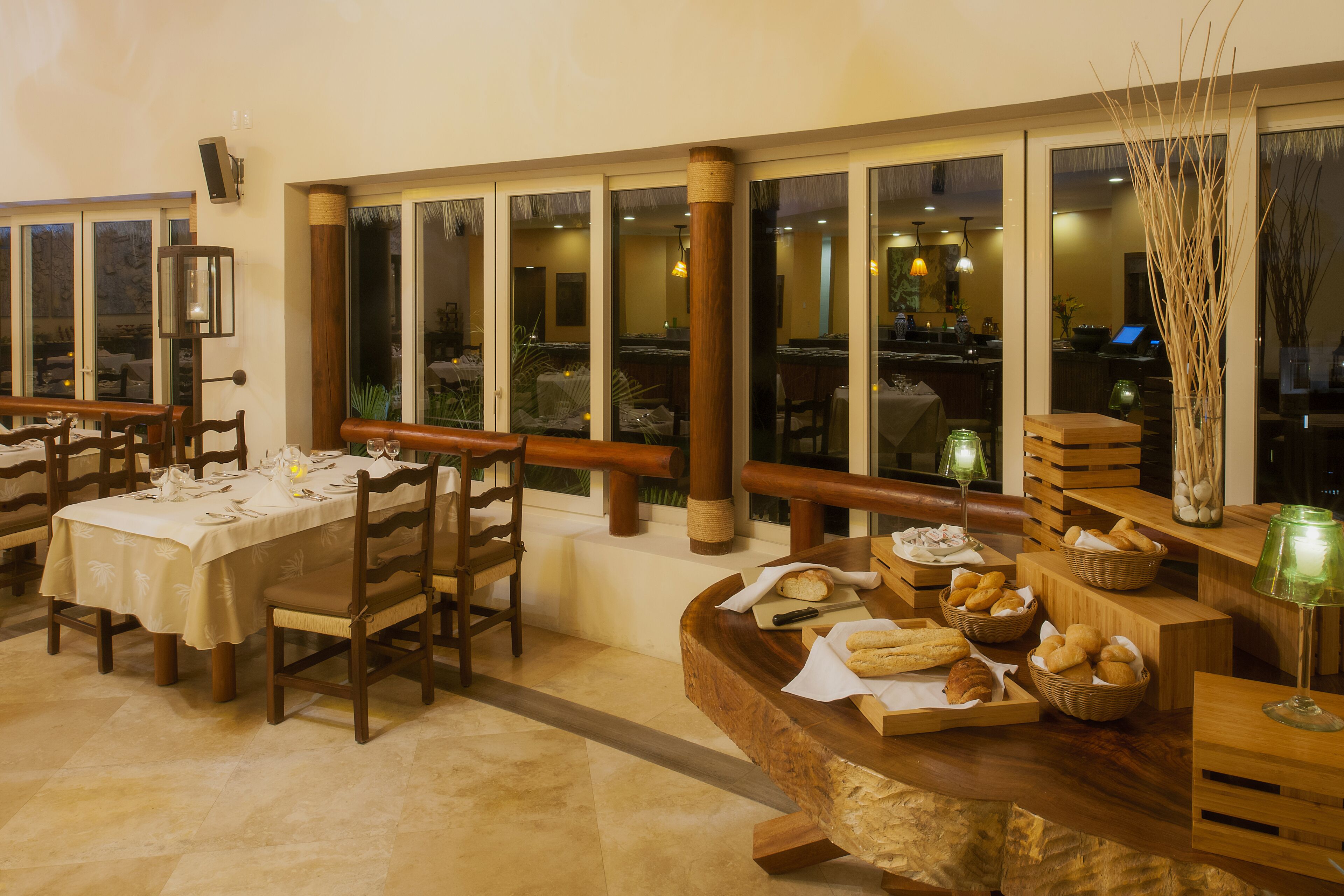 Een restaurant met houten tafels en stoelen, een buffet met brood en gebak, en grote ramen met uitzicht op de omgeving.