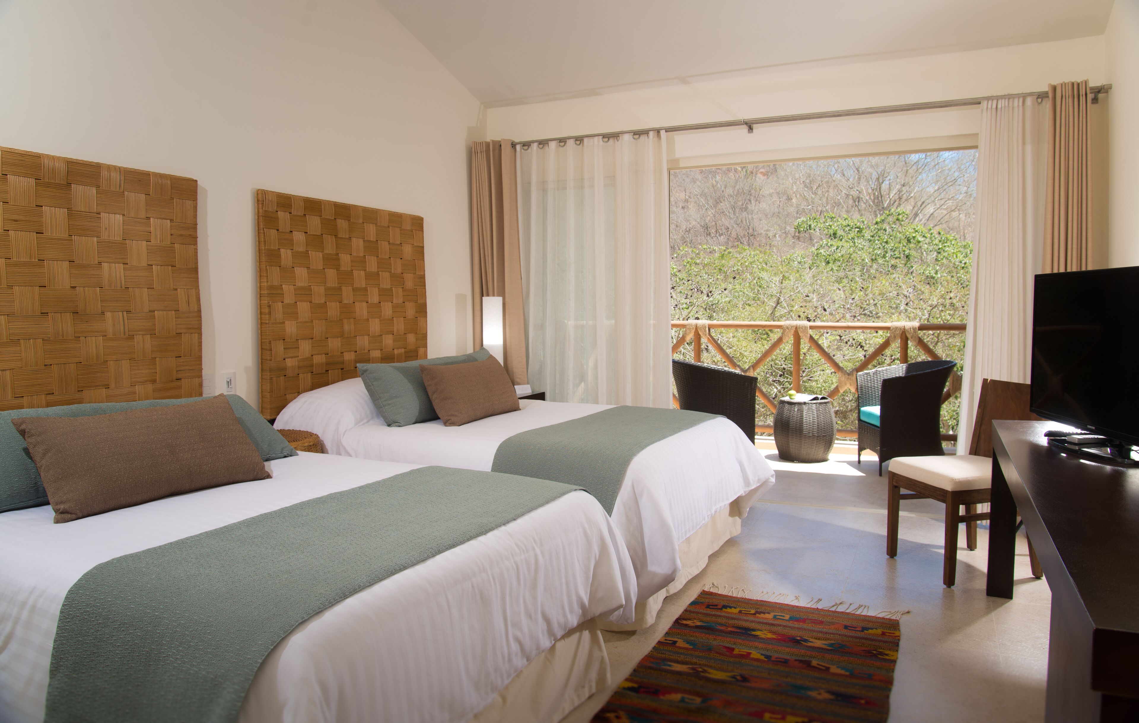 Habitación de hotel con dos camas, televisor, balcón con una silla y vista a árboles.