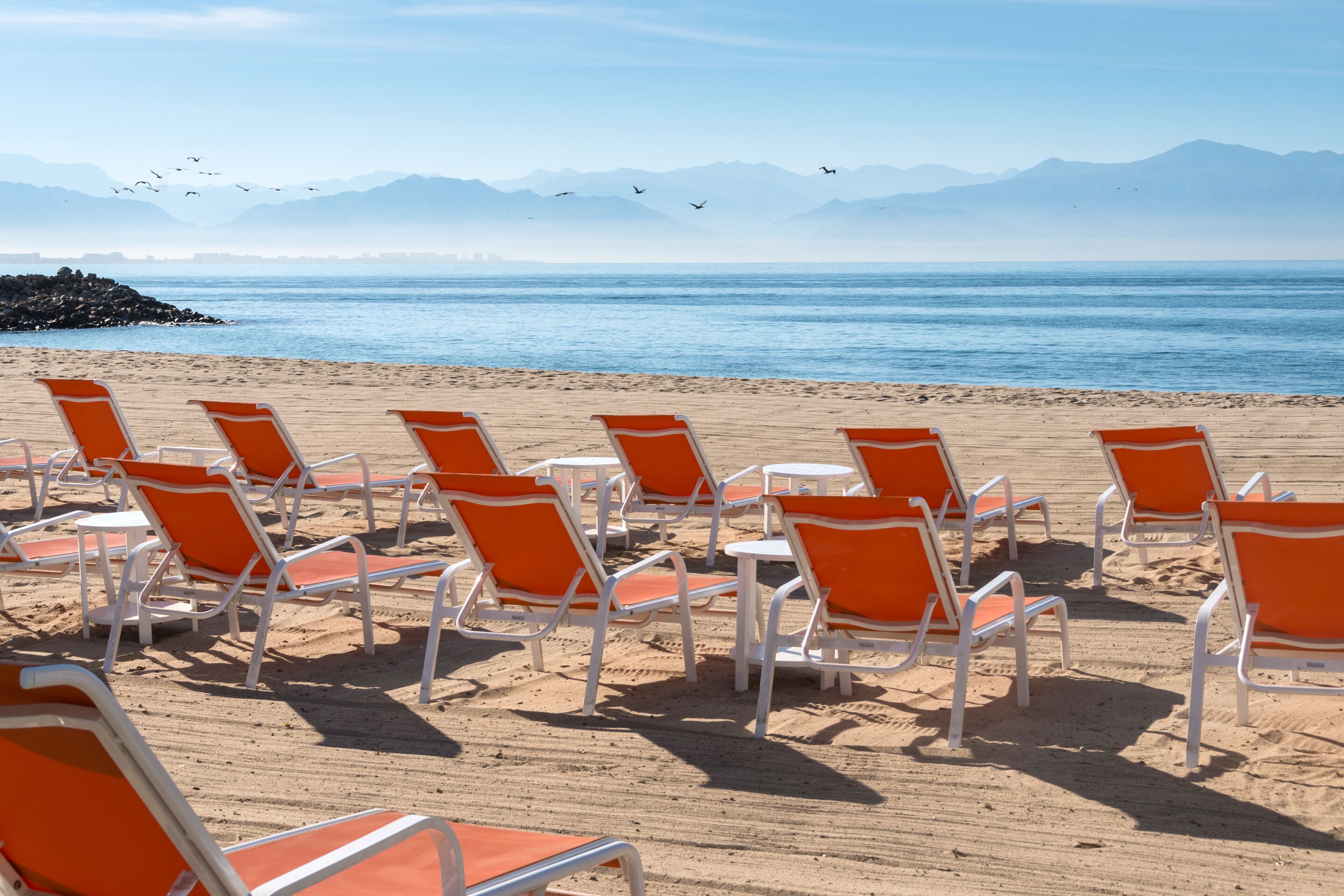 A beach with rows of orange and white beach chairs and tables, a clear view of the ocean, and mountains in the background.