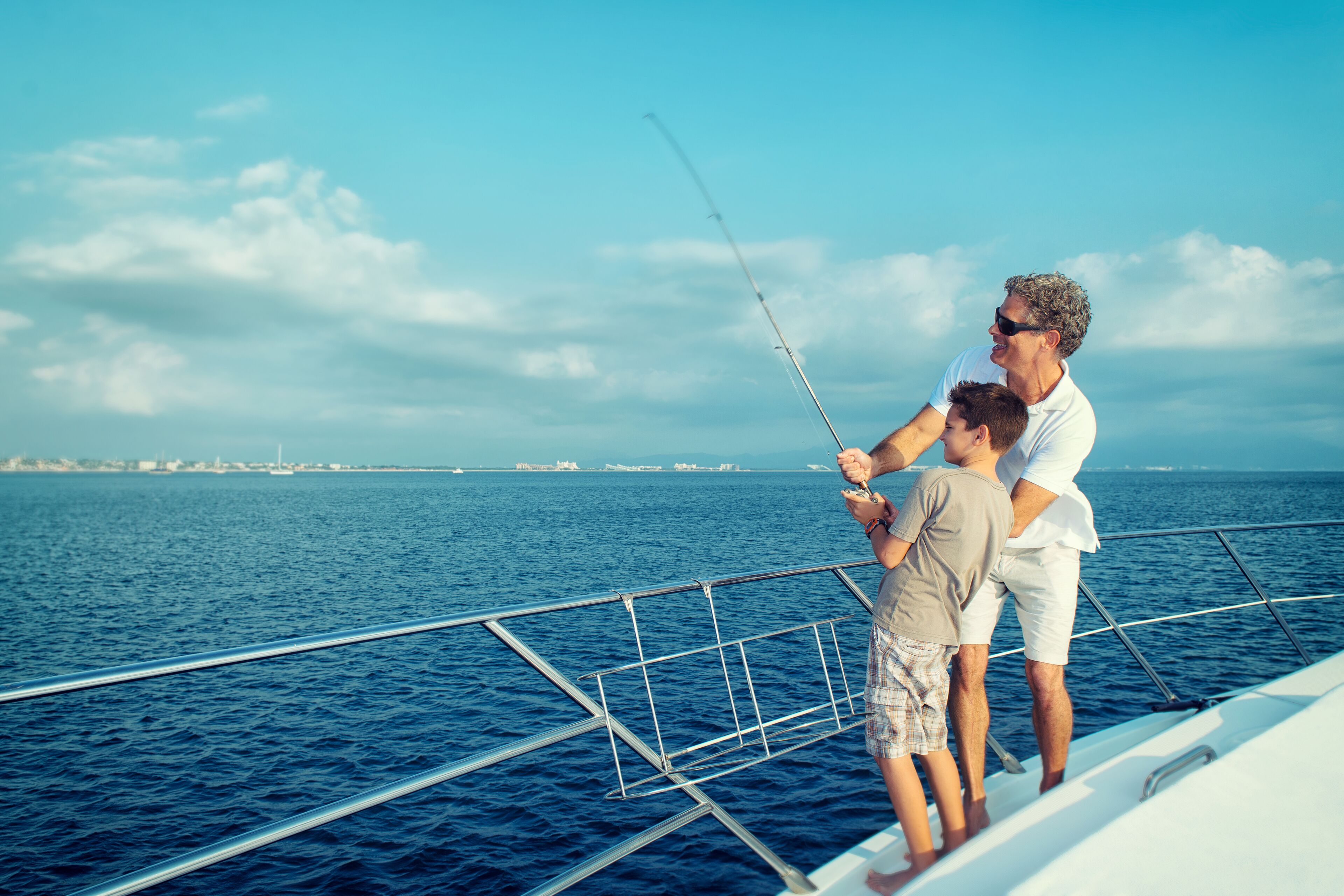 A man and a child fishing on a boat with a clear view of the ocean and distant shoreline.