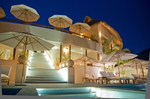 A pool area with lounge chairs and umbrellas, a staircase leading to a balcony, and a thatched roof structure.