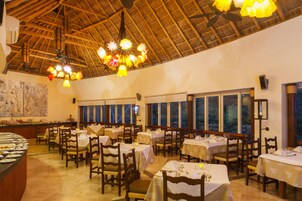 A restaurant with multiple tables and chairs, a buffet area, and a thatched roof ceiling.