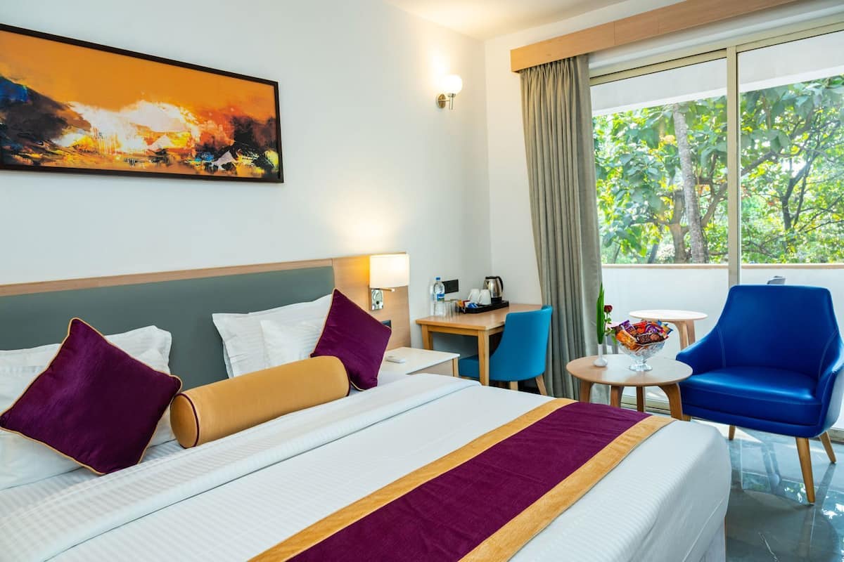 Une chambre d’hôtel avec un lit, un bureau, une chaise, une fenêtre donnant sur des arbres et un tableau au mur.