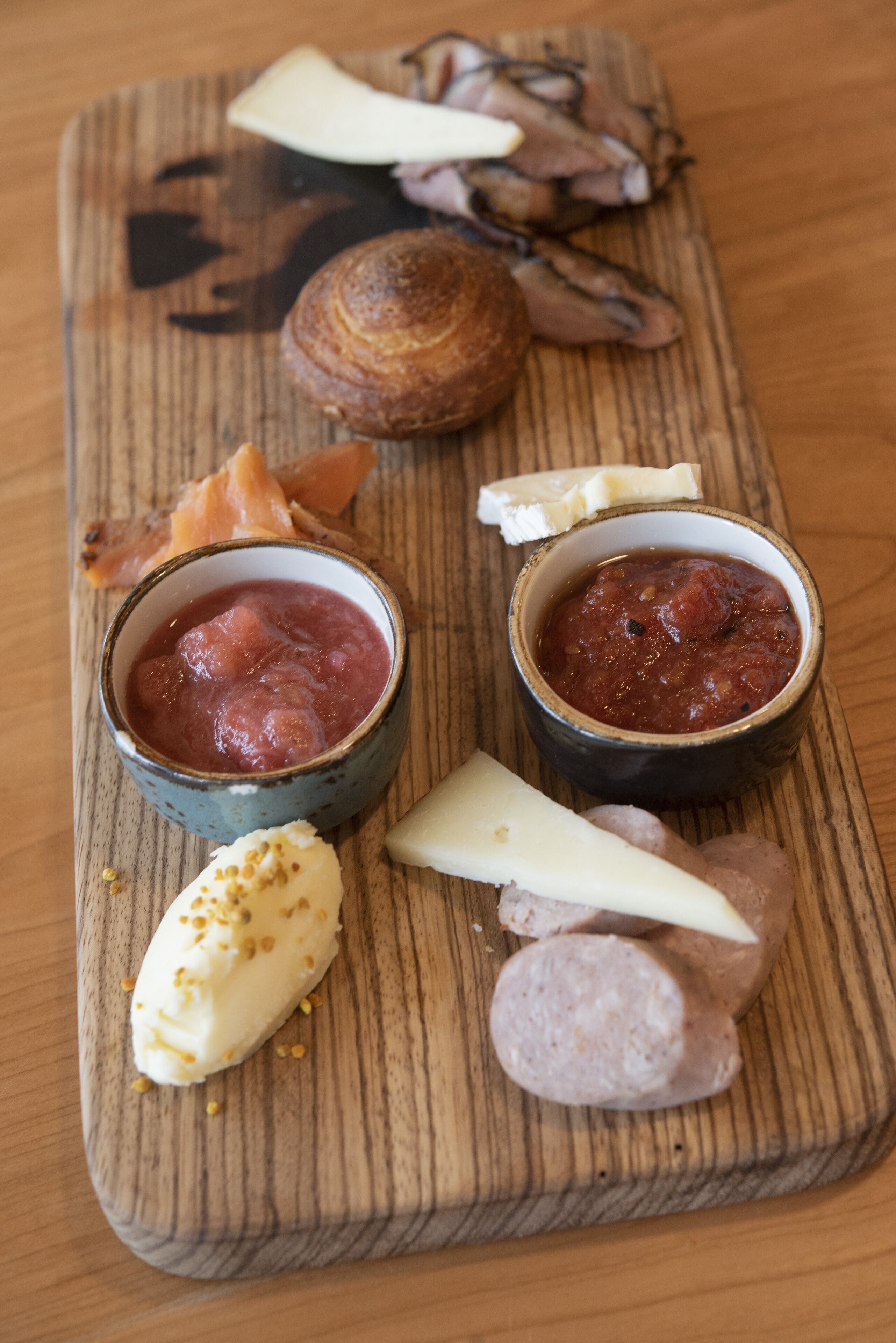 A wooden board with various food items including cheese, sausage, and a bowl of red sauce.