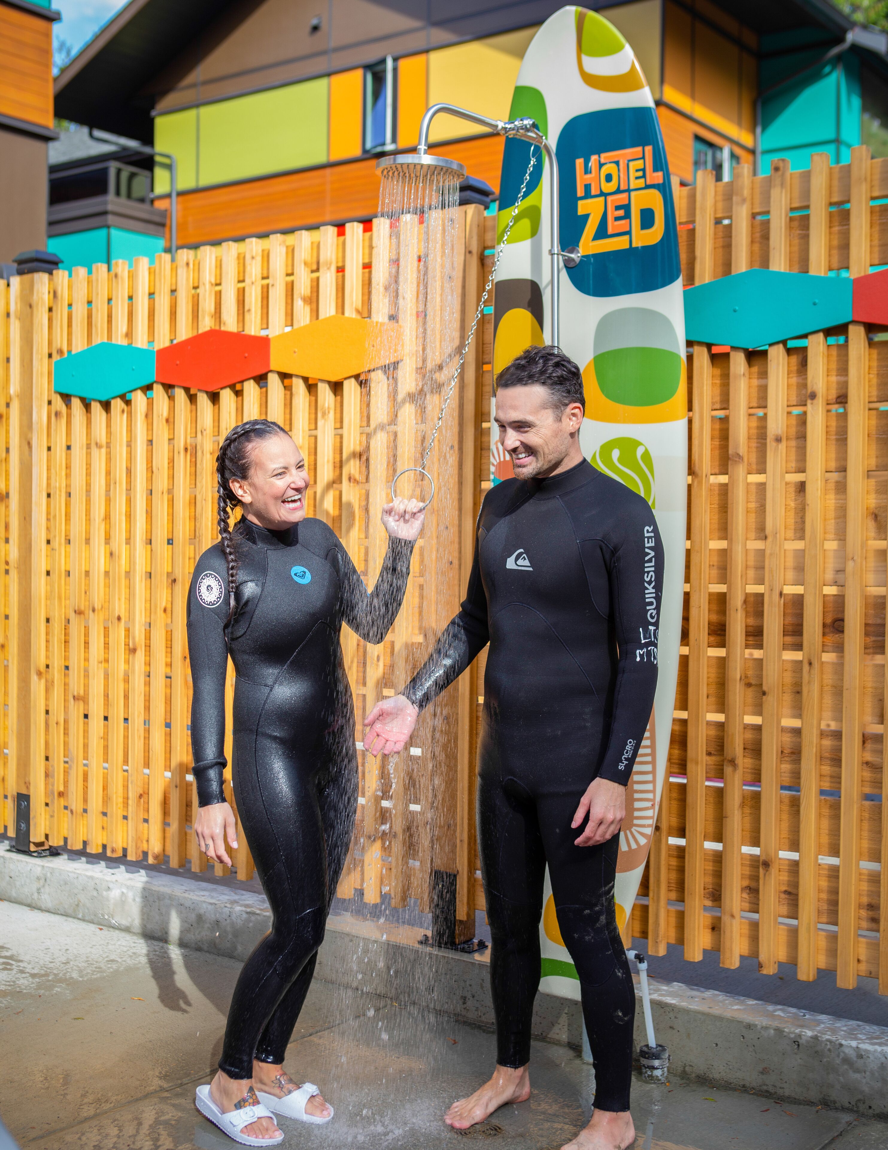 Two people in wetsuits standing in front of a wooden fence with a surfboard and a sign that reads "HOTEL ZED".