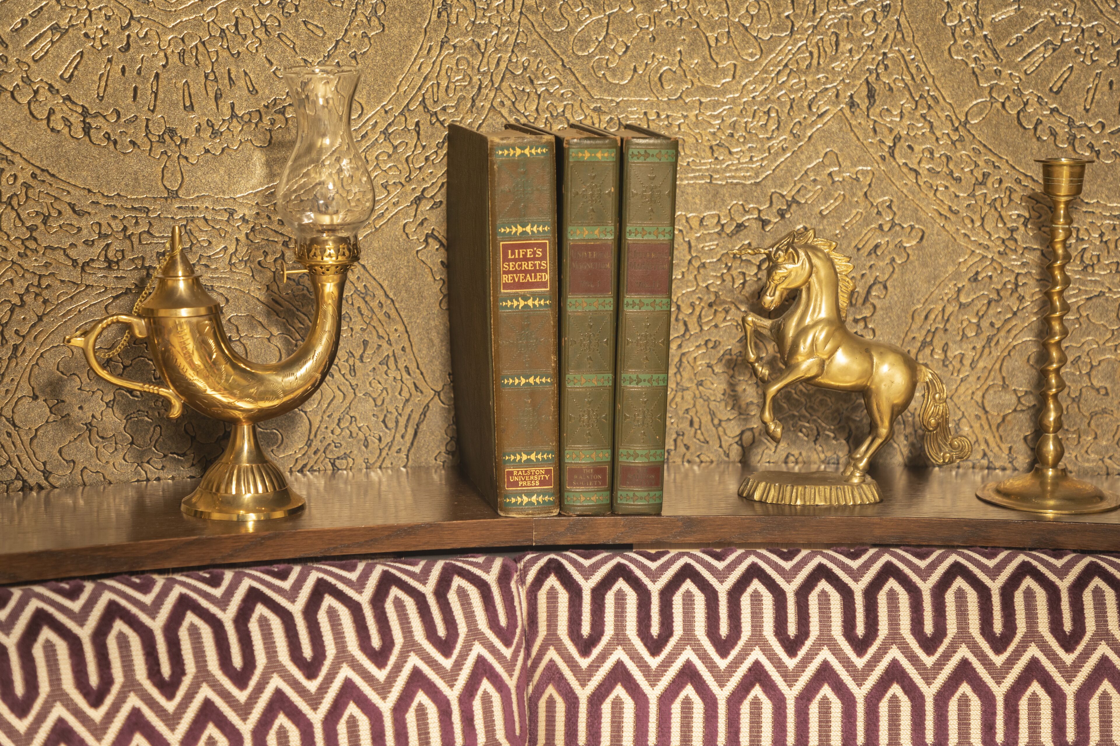A wooden shelf with three books titled 'Life's Secrets Revealed' by Bradford Press, a brass candlestick, and a brass horse figurine.
