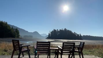 A wooden deck with four chairs and a table, overlooking a calm body of water and distant mountains under a clear sky.