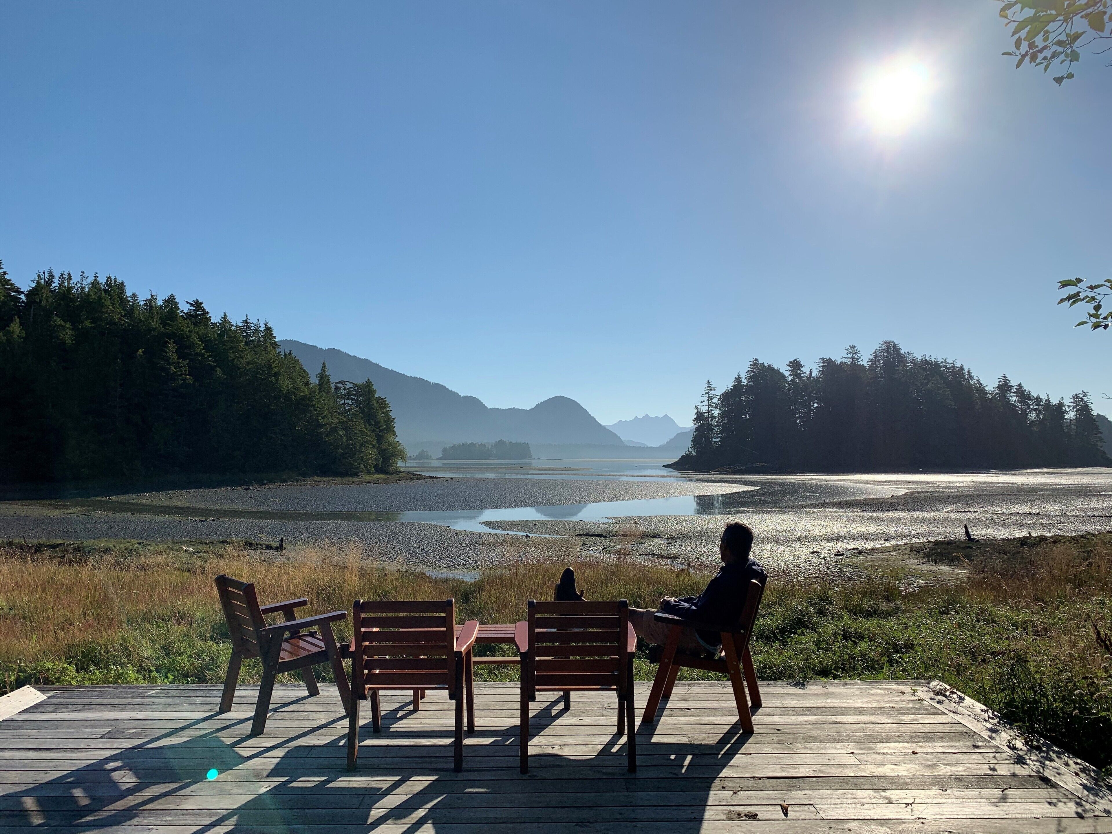A wooden deck with four chairs facing a calm river and distant mountains.