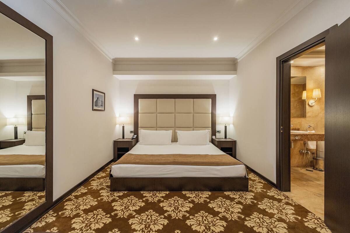 Ein Hotelzimmer mit einem großen Bett, Nachttischen inklusive Lampen, einem Spiegel und einem durchgegangenen Tür sichtbaren Badezimmer.