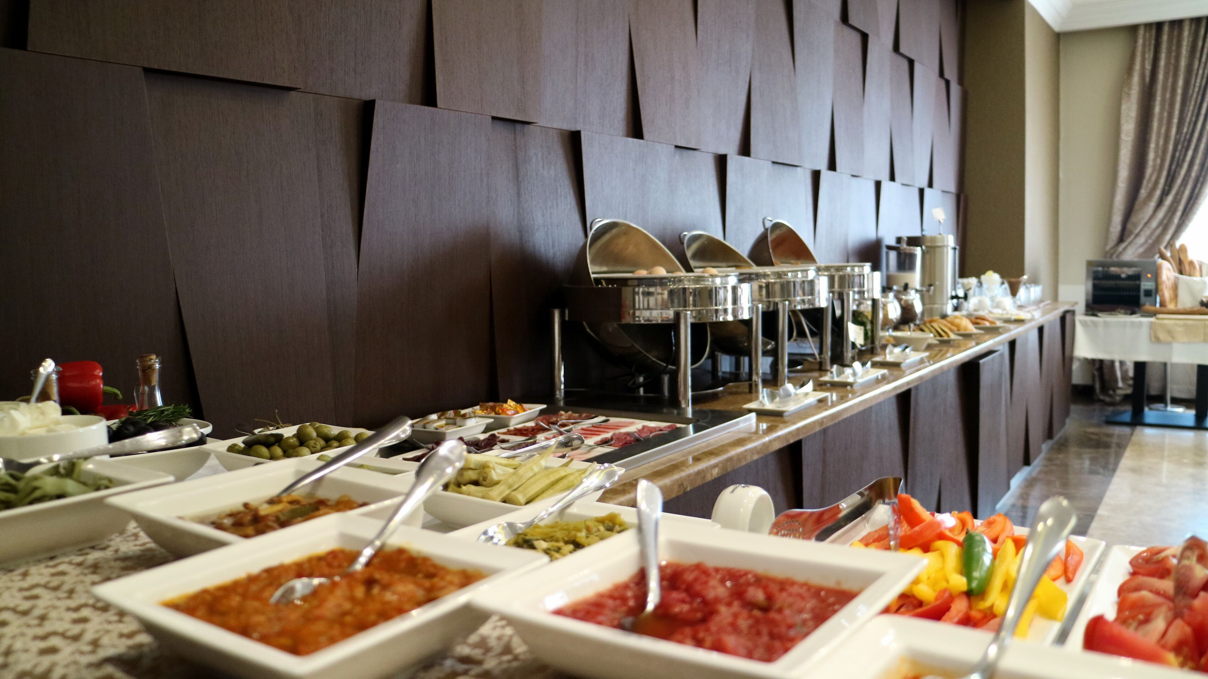 Un bufé con diversos alimentos y utensilios, dispuesto sobre una larga mesa frente a una pared con diseño geométrico.
