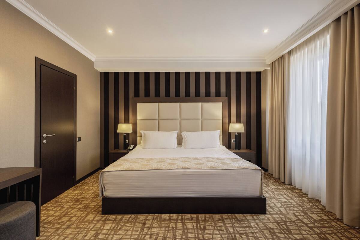 Ein Hotelzimmer mit einem großen Bett, zwei Nachttischlampen, einem dunkelholzigen Kopfteil, einem gemusterten Teppich und einer Tür links.