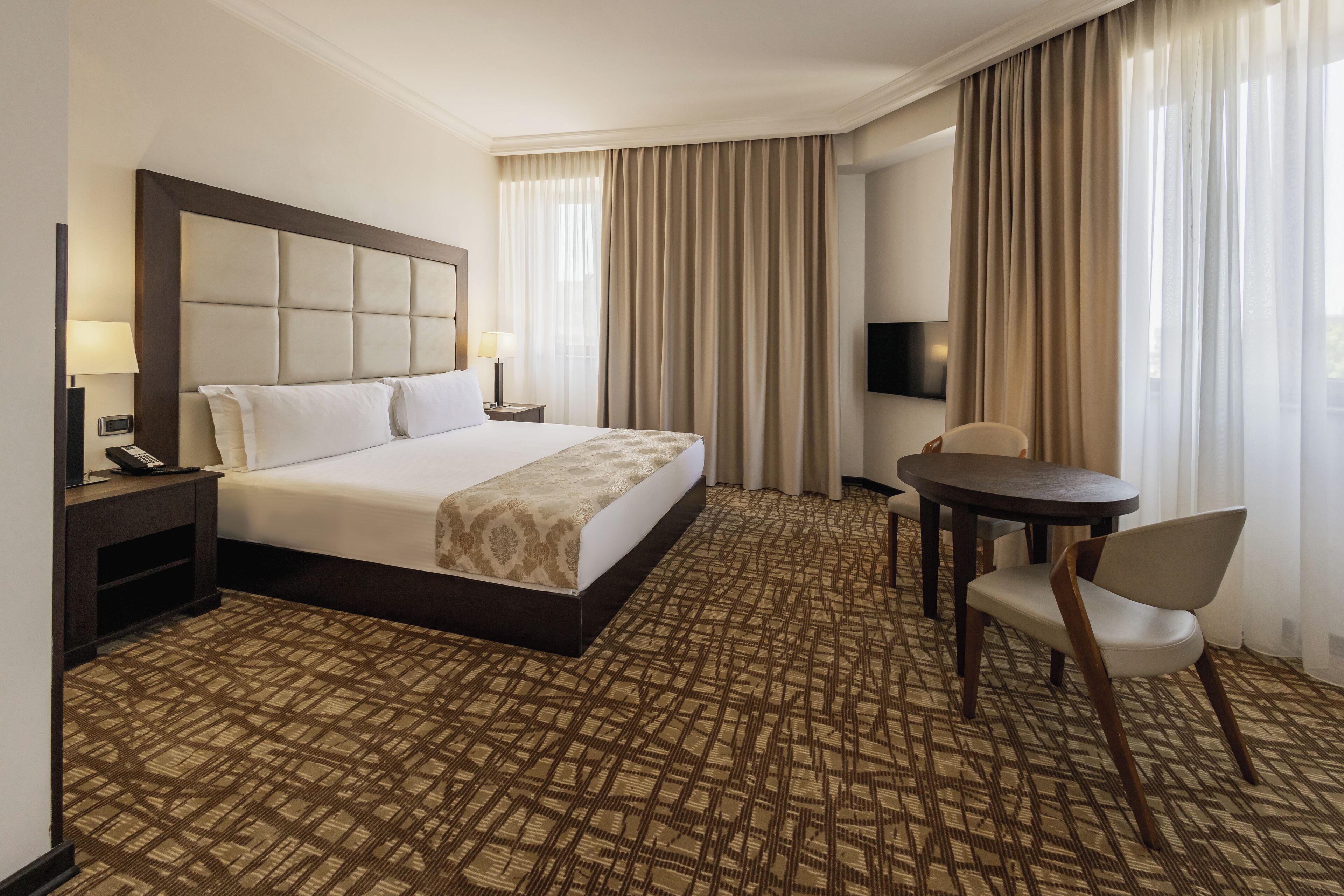 Una habitación de hotel con una cama grande, una mesita de noche con lámpara, una silla y una pequeña mesa.