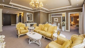 Een ruime woonkamer met een grote gouden zithoek, een witte salontafel en een kroonluchter.
