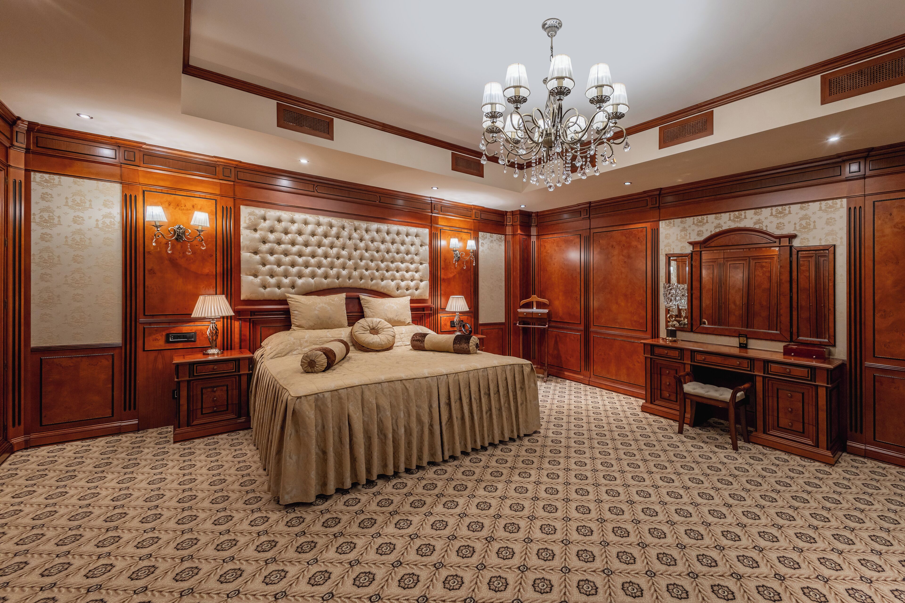 A luxurious bedroom with a large bed, a chandelier, wooden paneling, and a patterned carpet.