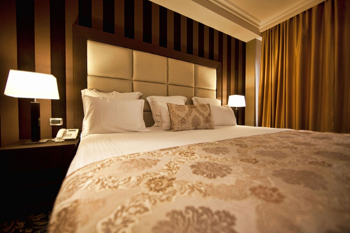 Una habitación de hotel con una cama grande, un cabecero con listones verticales, dos lámparas de noche y un teléfono en la mesita de noche.