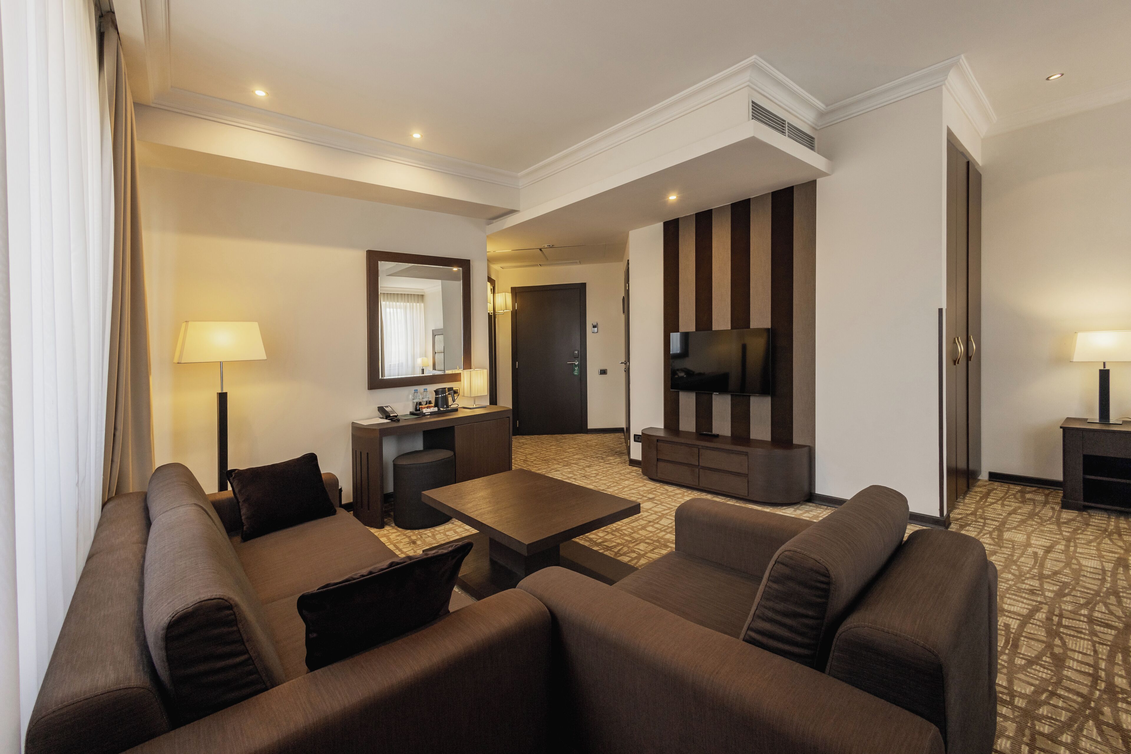 A modern hotel room with a living area featuring a sofa set, a coffee table, a TV, and a small kitchenette.