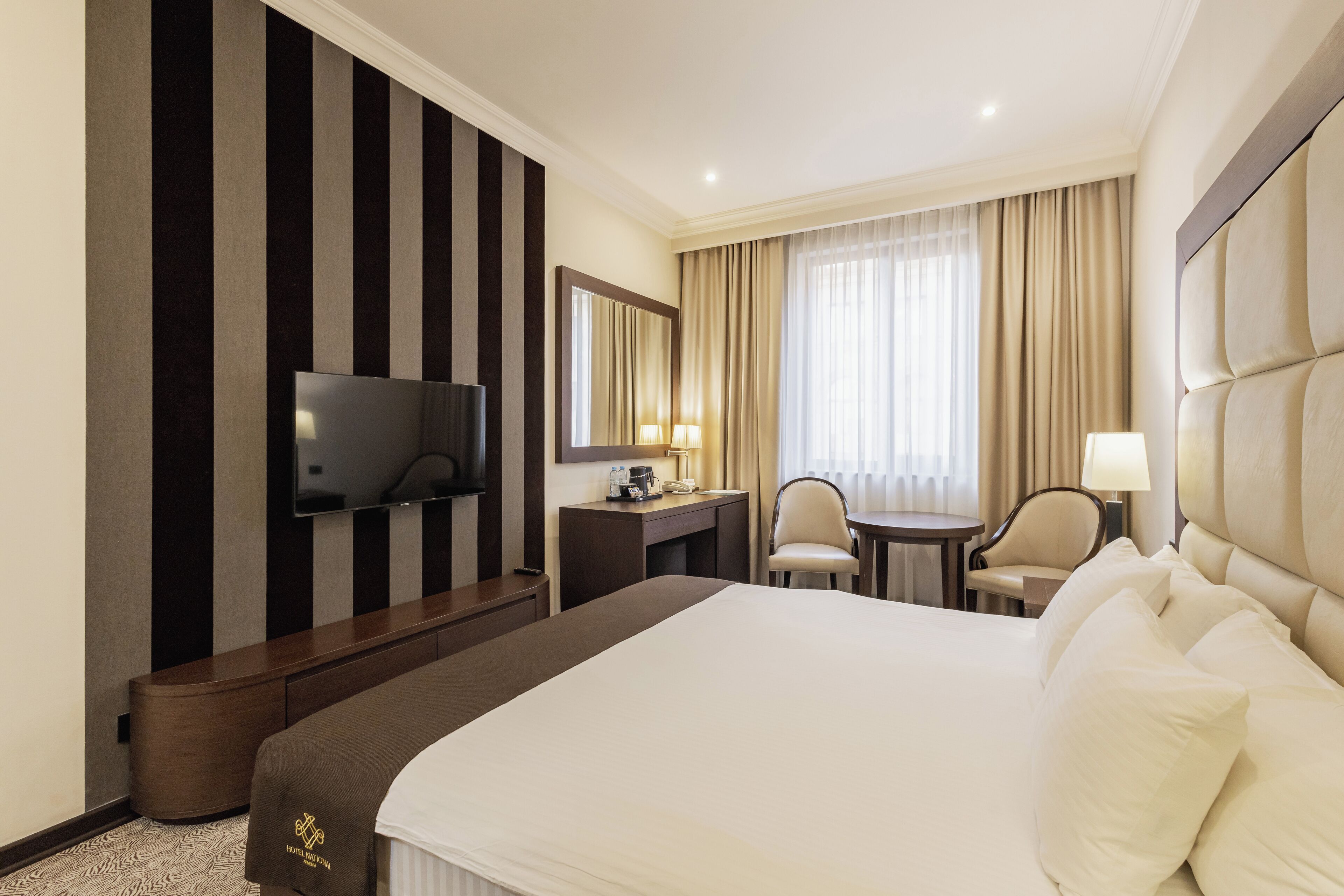 A hotel room with a large bed, a desk with a flat-screen TV, a chair, and a small table.