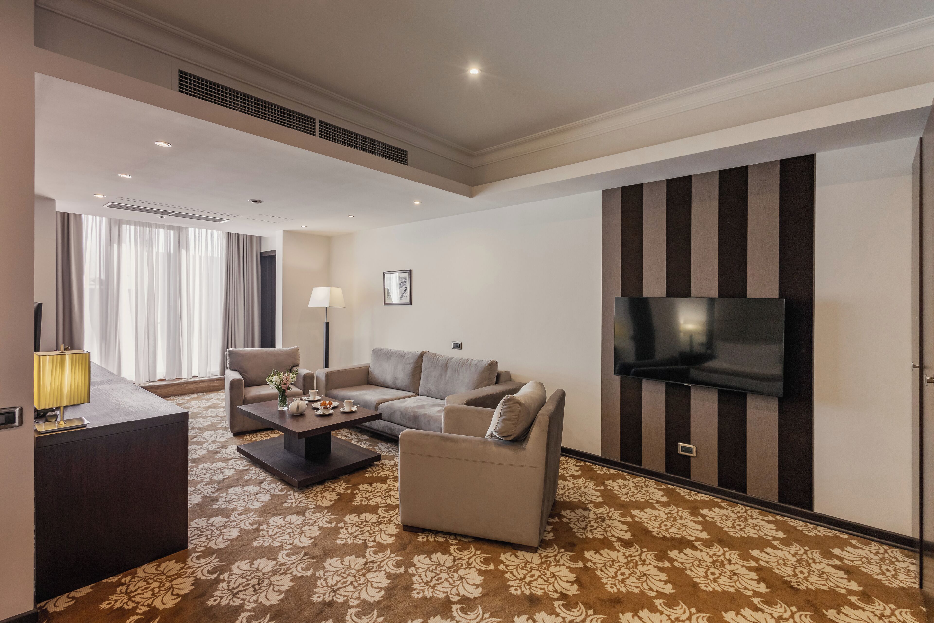 Una habitación de hotel moderna con un conjunto de sofás, un televisor de pantalla plana, un escritorio con lámpara y una alfombra estampada.