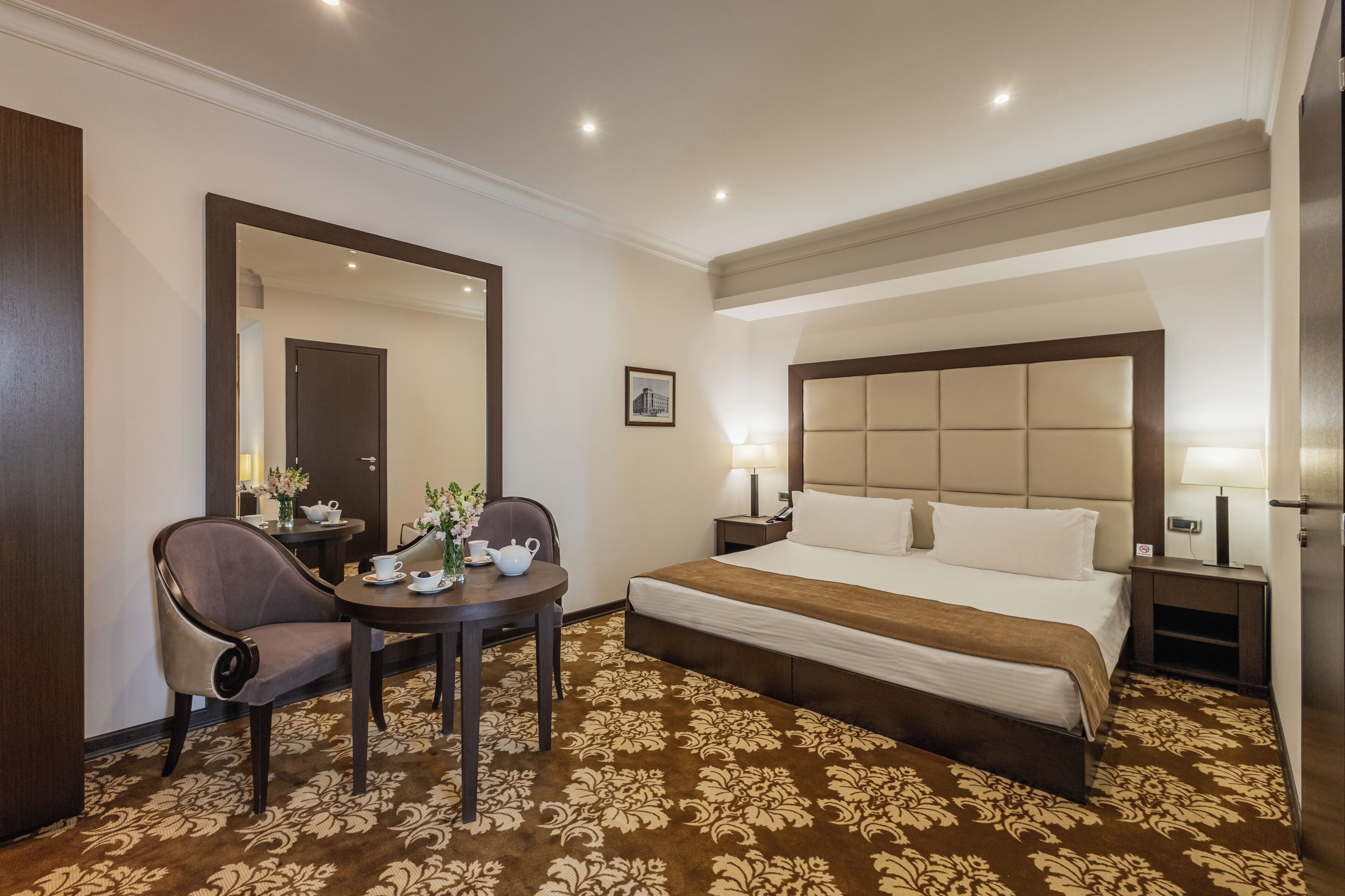 Una habitación de hotel con una cama grande, un área de descanso con una mesa y sillas, un espejo y una mesita de noche con lámpara.