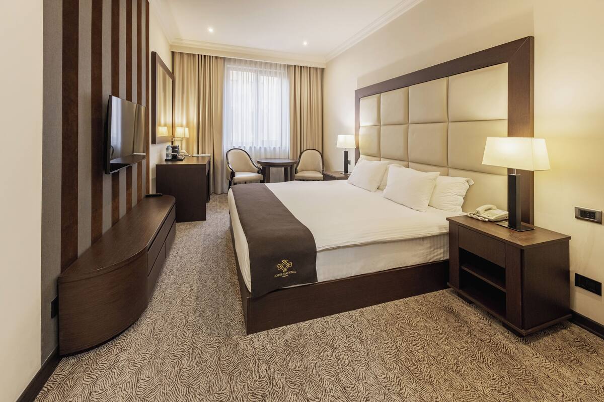 Ein Hotelzimmer mit einem großen Bett, einem Fernseher, einem Schreibtisch mit Lampe und einem Sessel.