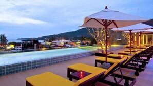 Un área junto a la piscina con sillones de descanso y sombrillas, una piscina con azulejos y vistas a un pueblo y montañas al fondo.