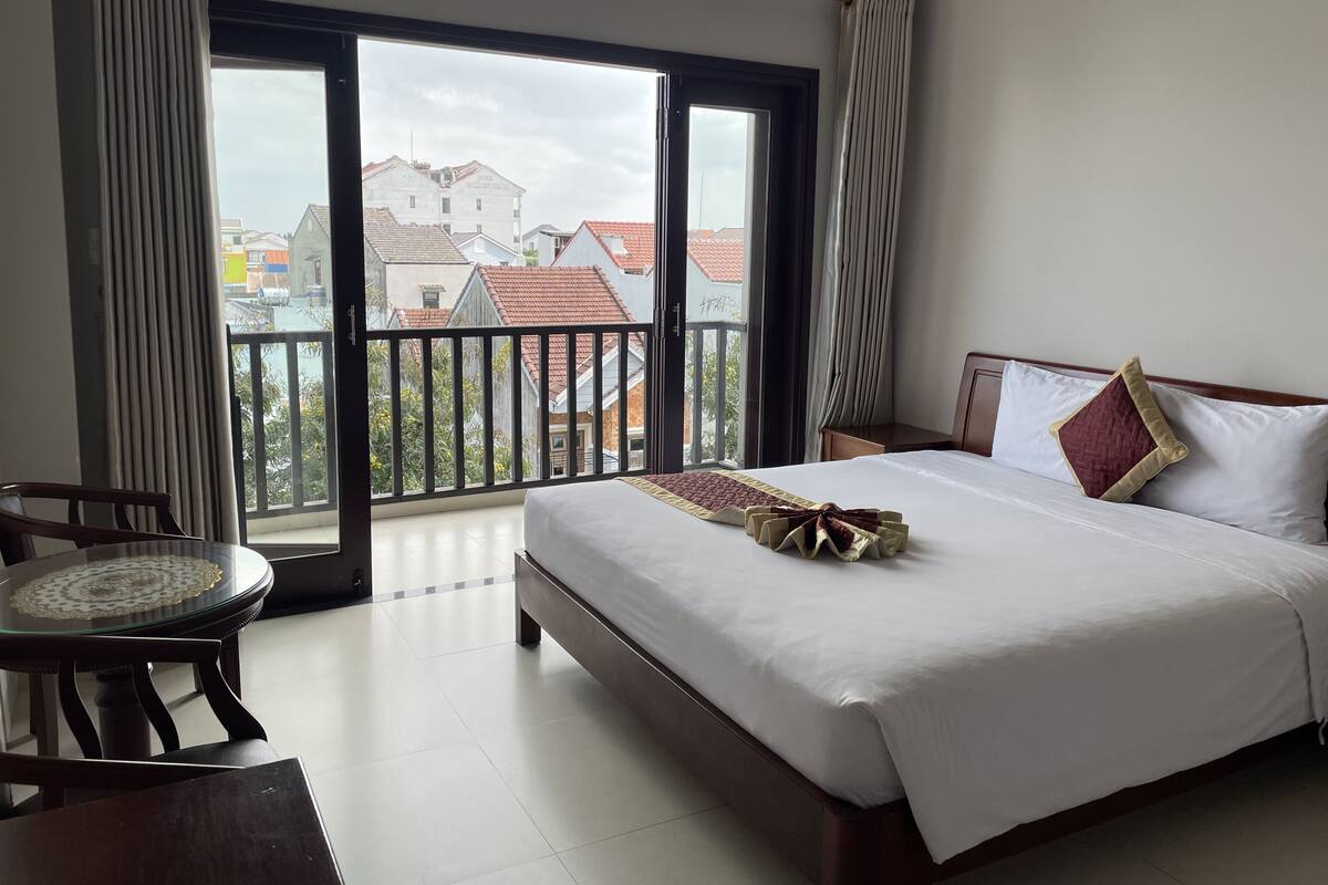 A hotel room with a large bed, a round table with chairs, and a balcony with a view of buildings.