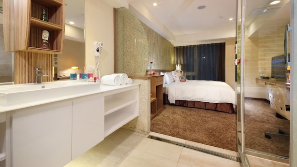 A hotel room with a large mirror, a wooden vanity, a bed with a brown carpet, and a glass partition. - Nagahiro Hotel (Taipei)