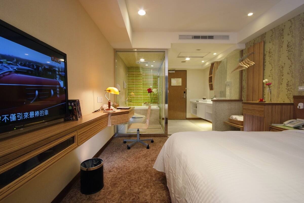 A hotel room with a bed, a desk with a chair, a television, a mirror, and a bathroom visible through a glass partition.