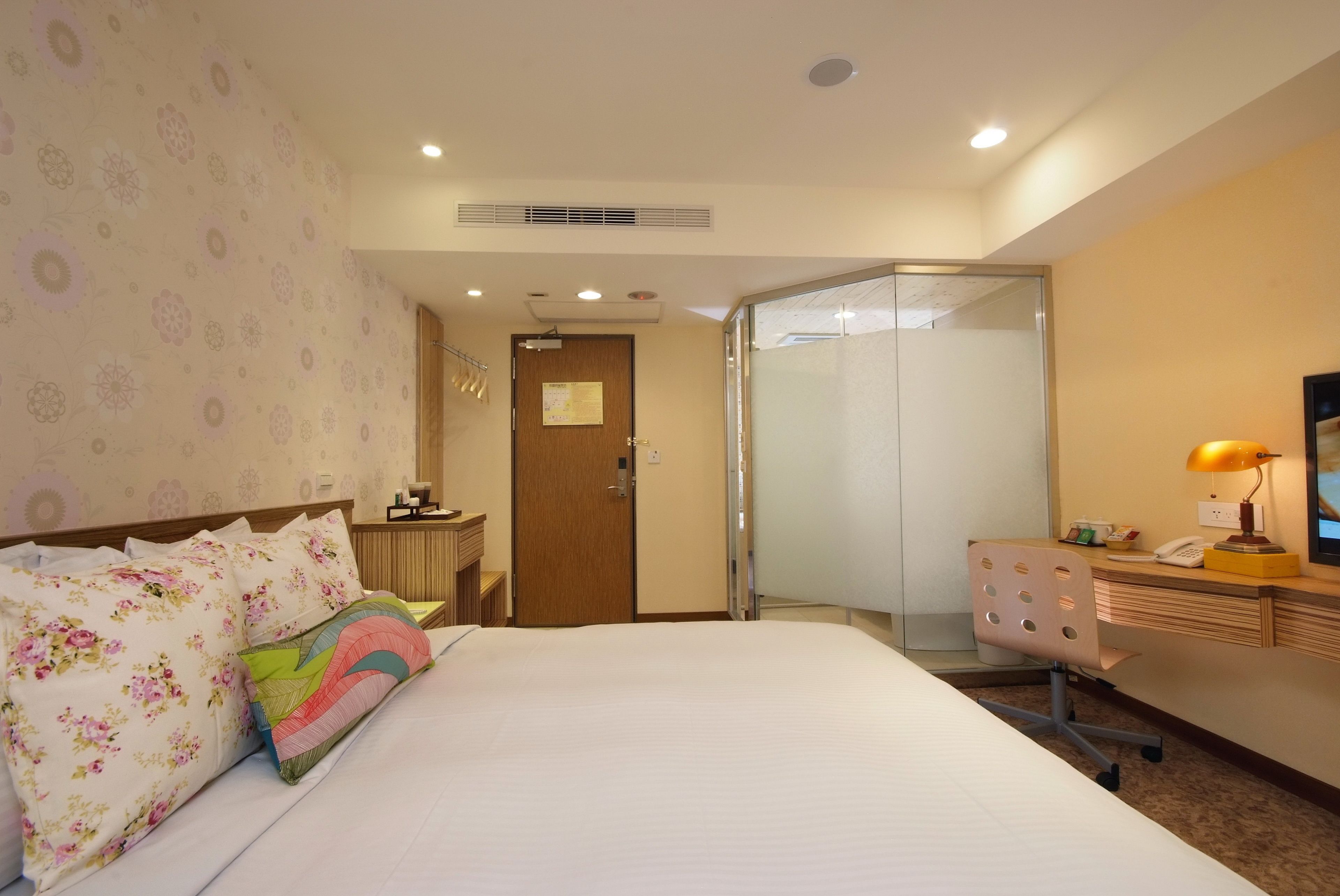 A hotel room with a bed, a desk, a chair, a lamp, and a door leading to a bathroom.