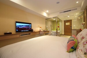 Een hotelkamer met een groot bed, een aan de muur bevestigde televisie, een badkamer met een douchecabine van glas, en een klein bureau met een lamp.
