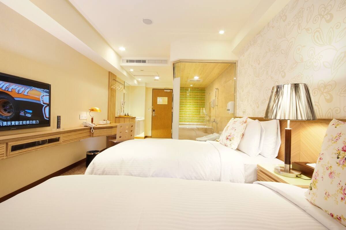 A hotel room with two beds, a TV, a desk with a lamp, and a shower area.