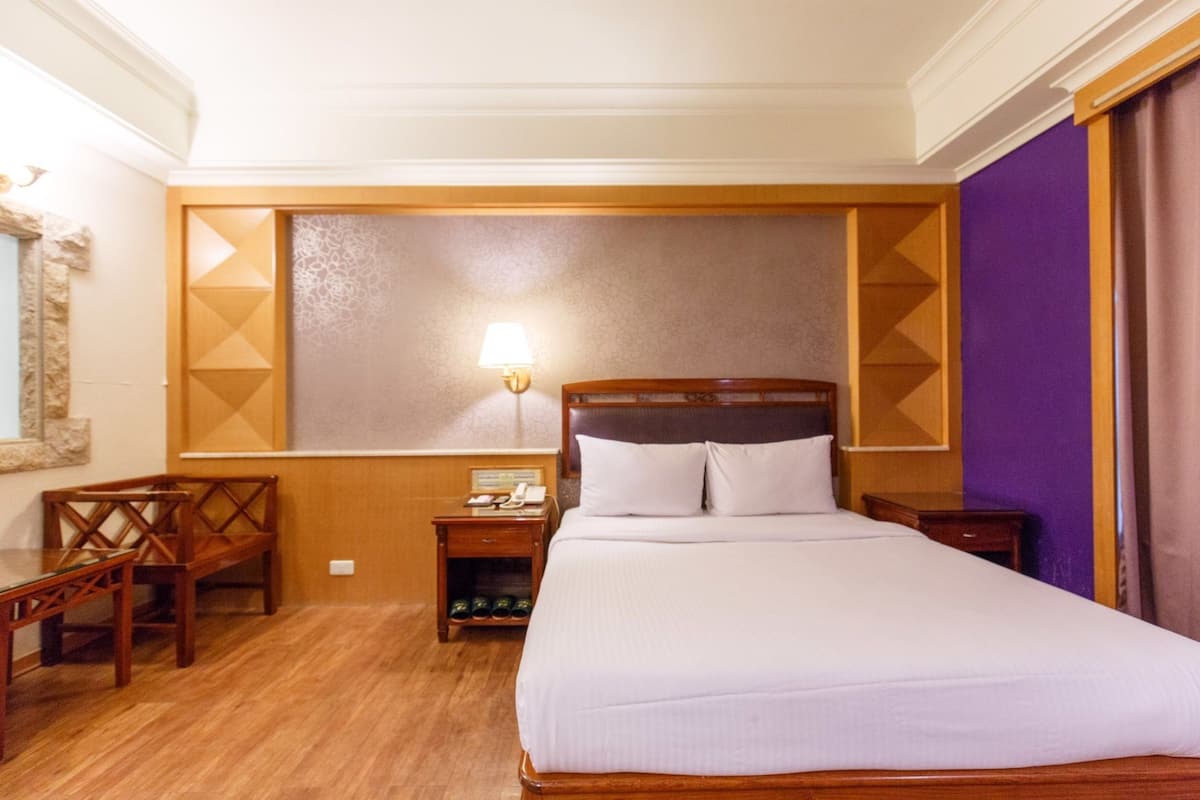 A hotel room with a large bed, wooden headboard, a nightstand with a lamp, a wooden chair, and a small table with a telephone.