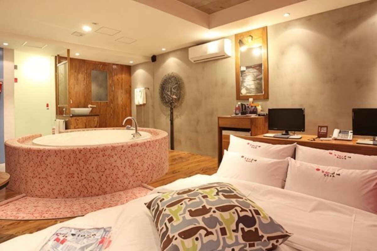 Une chambre avec une grande baignoire ronde, un lit agrémenté d’oreillers à motifs, une table de chevet avec une lampe et un téléviseur fixé au mur.