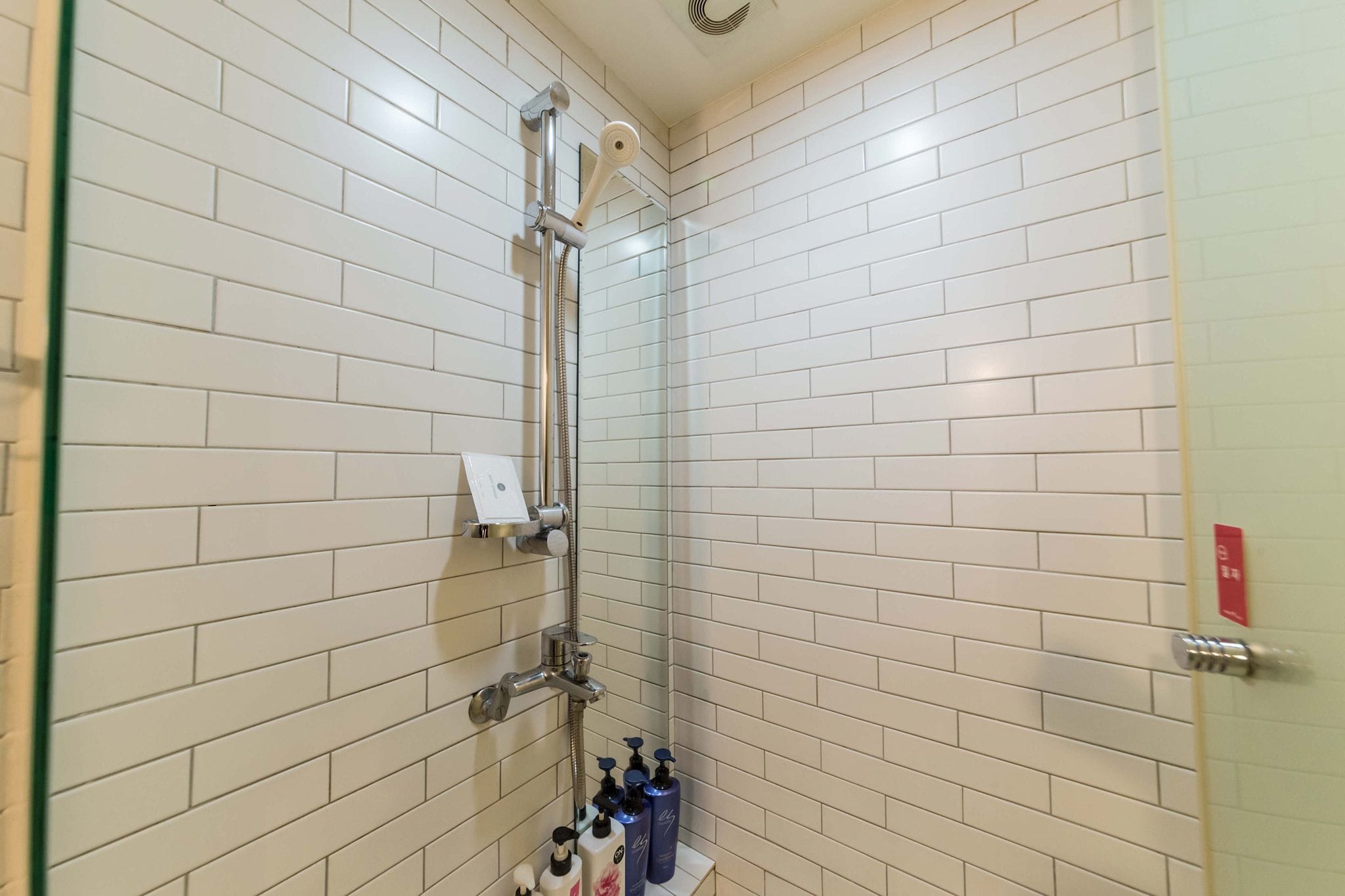 Un espace douche avec des murs recouverts de carreaux blancs, une porte en verre et une étagère contenant des articles de toilette.