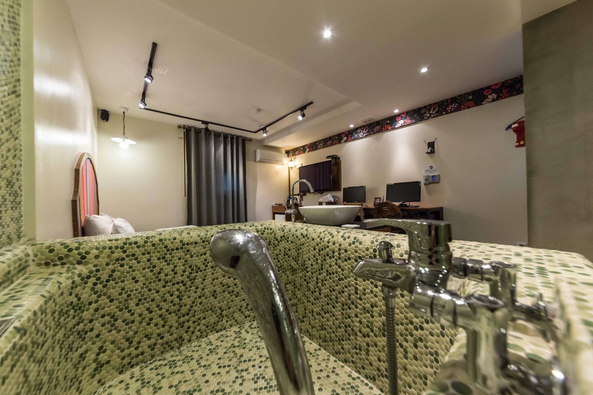 A bathroom with a large bathtub, a sink, and a mirror. There is a window with curtains, a towel rack, and a showerhead.