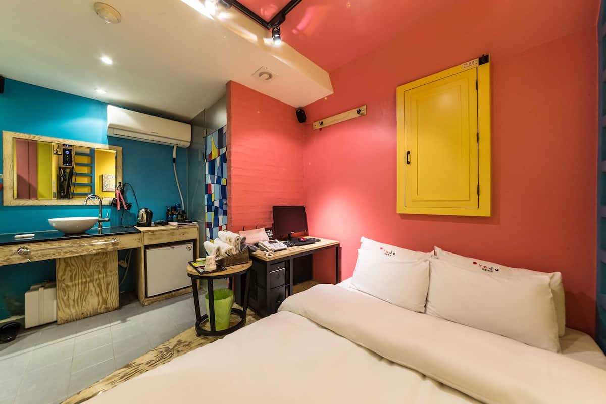 A bedroom with a bed, a desk, a chair, a small table, a mirror, and a wardrobe.
