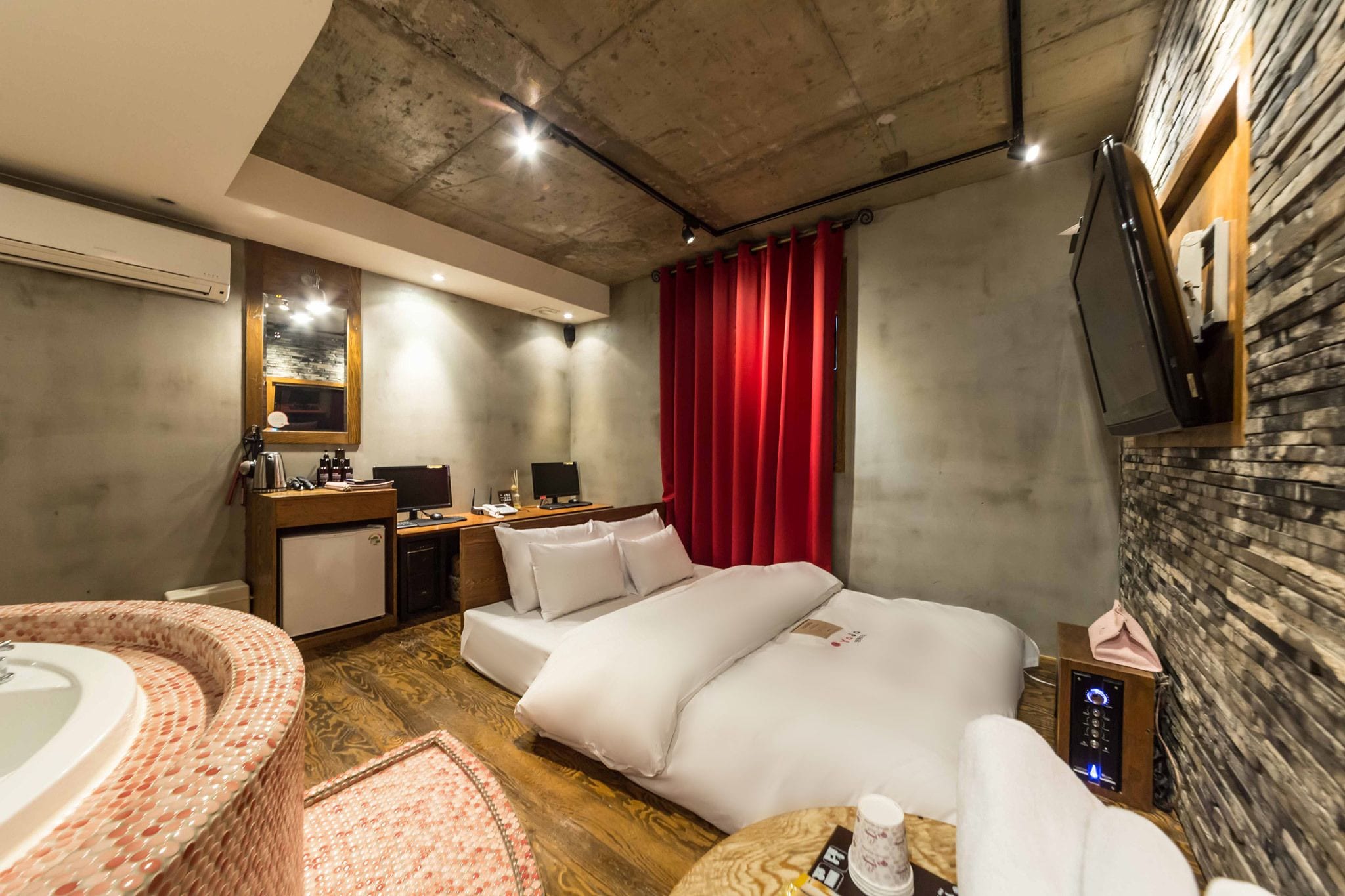 Una camera d'albergo con un letto, una scrivania, un angolo cottura, una televisione, una tenda rossa e un bagno con vasca e specchio.